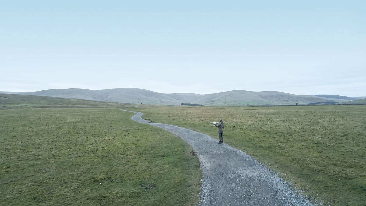 Sentiero di ghiaia in un prato verso colline lontane, con una piccola figura e un drone