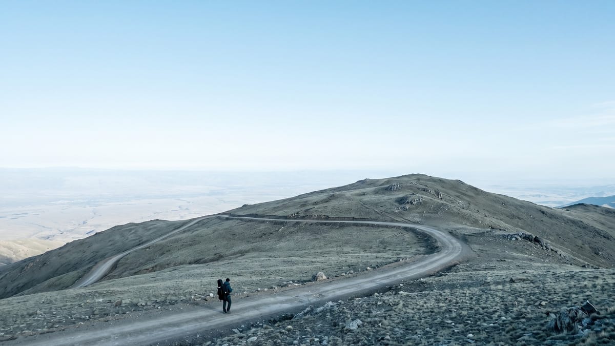 Strada di montagna a tornanti su un crinale, con una figura minuscola che sale con una valigia